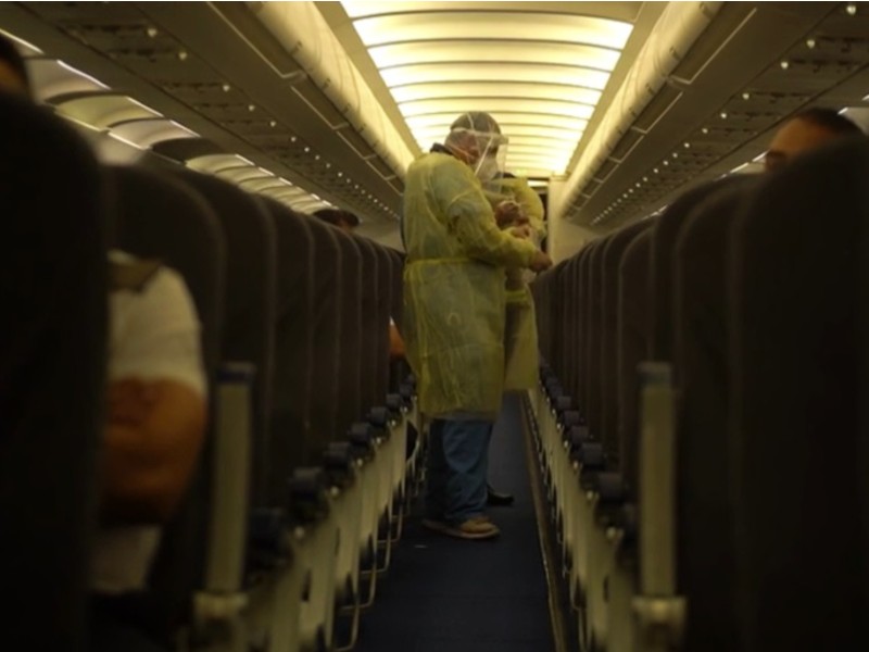 A imagem mostra a parte interior de um avião, com o corredor no centro e assentos pretos nas laterais da foto, chão azul escuro, luzes nos compartimentos de malas e no teto. Em um dos assentos aparece o braço de uma pessoa que veste uma camisa branca. No corredor, um homem com calça azul, tênis cinza, avental amarelo e face shield cobrindo o rosto está colocando luvas nas mãos
